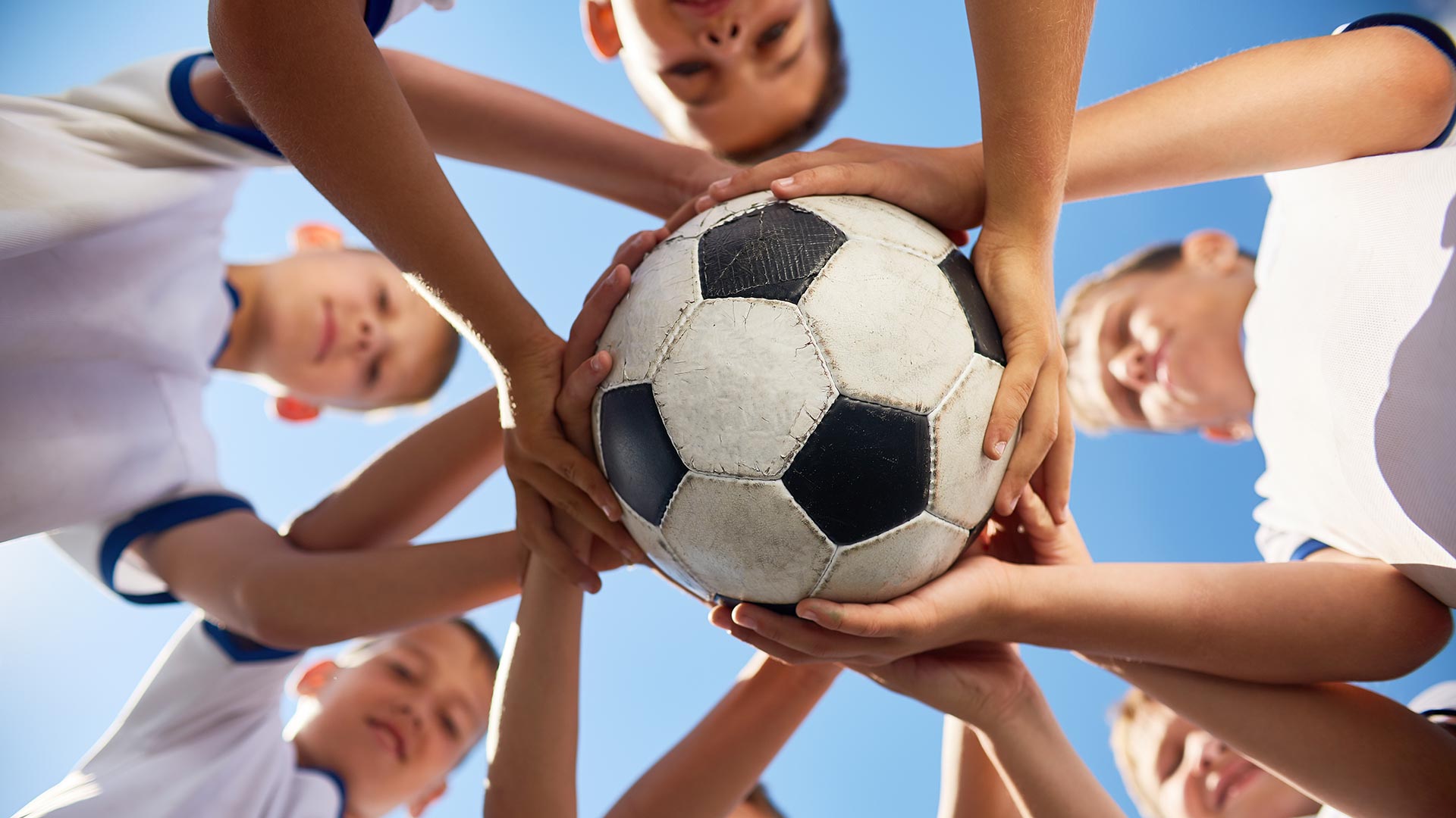 Low angle view of boys in junior football team standing in circle holding ball together against blue sky, focus on ball