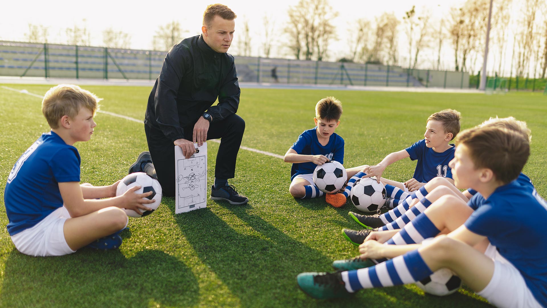 Group of Young Boys Sitting on Sports Grass Field witch School Coach. Kids Listening Coach's Tactic Talk. Young Coach Explain Football Tactic. Coaching Youths in Sports